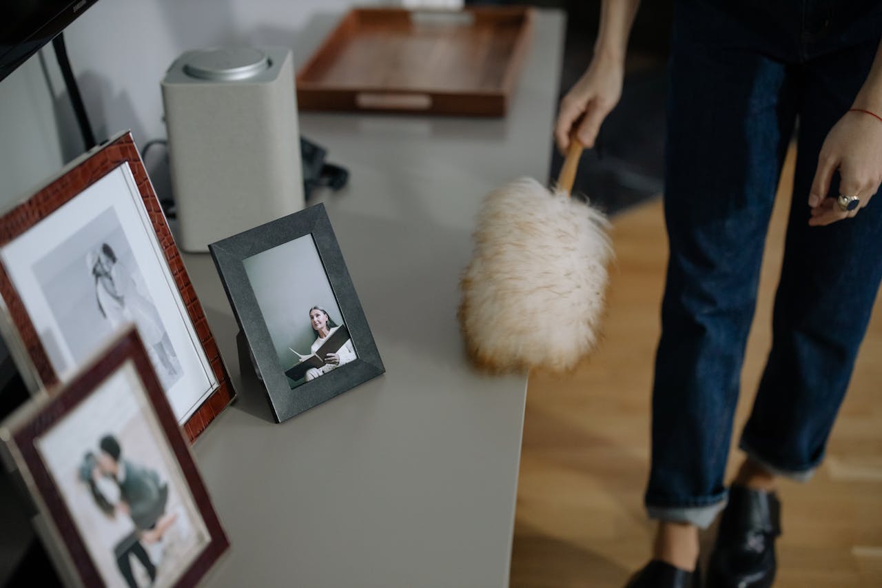 A woman using a feather duster to clean picture frames on shelves indoors.