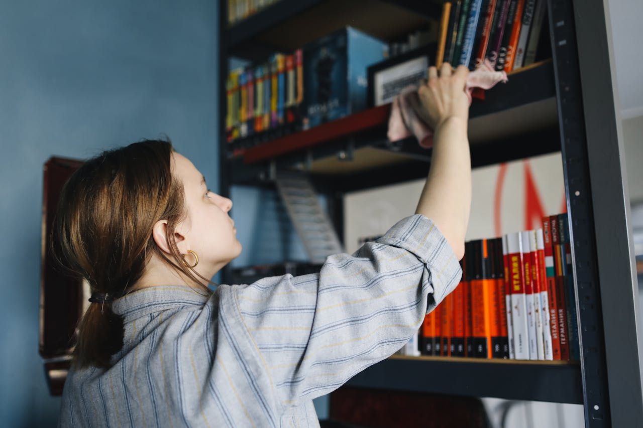 A woman cleans a bookshelf using a cloth, organizing books in a tidy home environment.